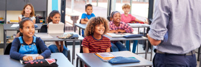 Students smiling in a classroom while a teacher is going over a lesson.